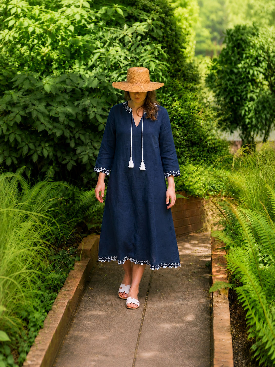 Woman wearing a navy linen dress with white embroidery and tassel ties in a garden setting by Kaminee London
