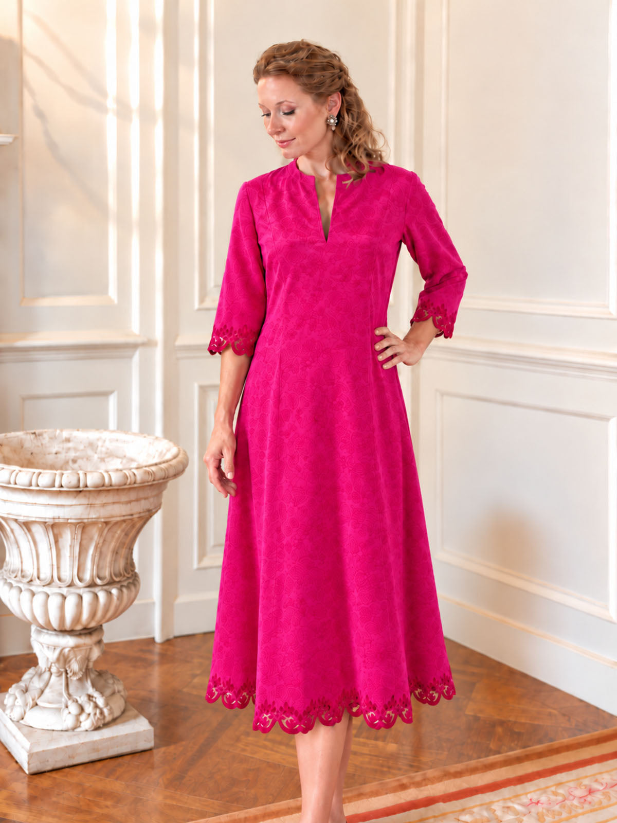 A woman wearing a bright fuchsia pink midi dress with three-quarter sleeves and scalloped lace trim, standing in a classic white-paneled room with a wooden floor and a decorative stone planter.